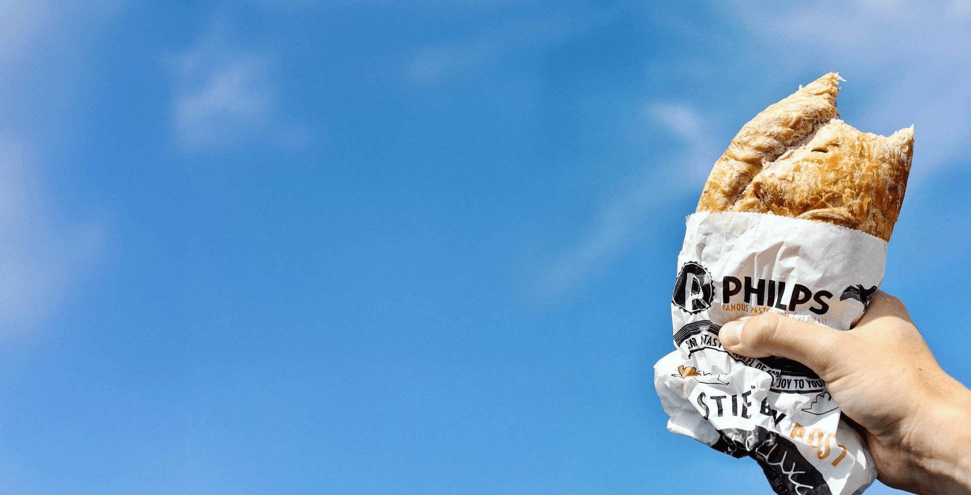 Hand holding a Philps-wrapped Cornish pasty against a bright blue Cornish sky